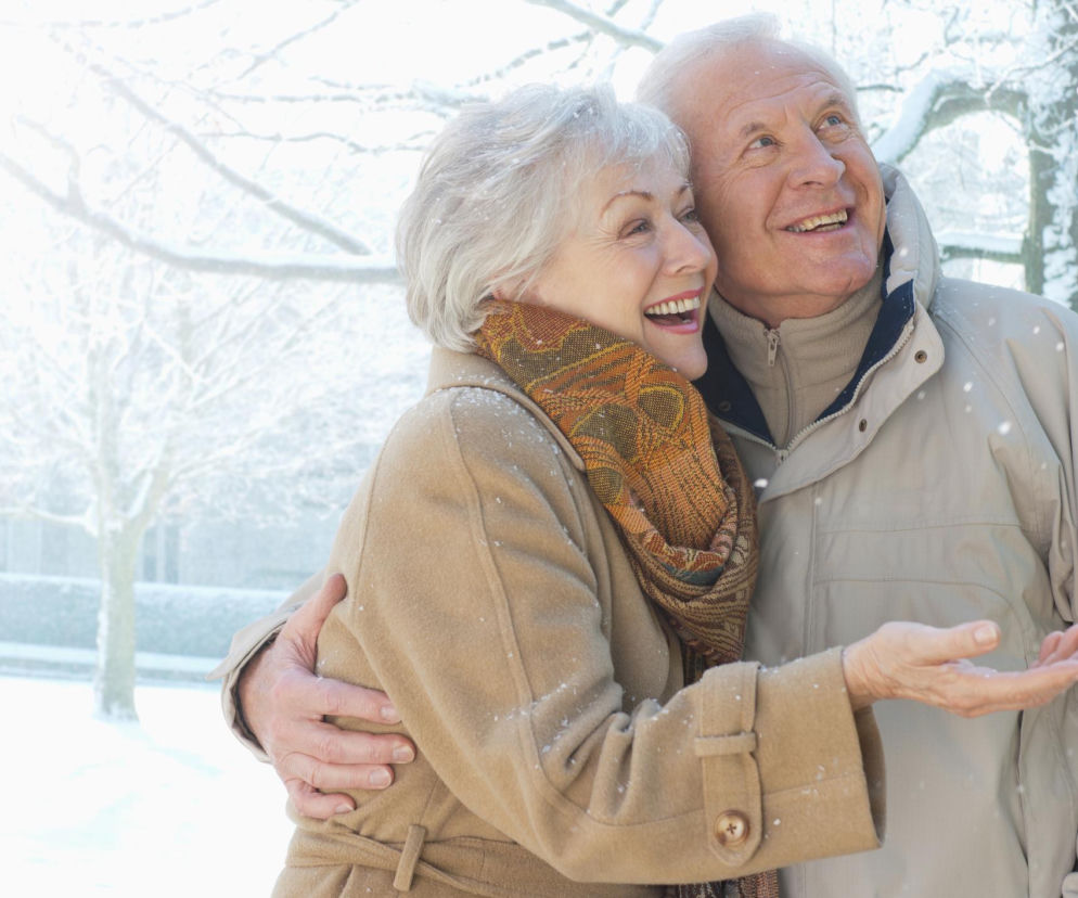 Uśmiechnięta para starszych osób w zimowych ubraniach, otoczona padającym śniegiem i ośnieżonymi drzewami. Mężczyzna obejmuje kobietę, a oboje patrzą w górę, jakby podziwiali płatki śniegu. Scena symbolizuje radość z zimowego wypoczynku, o którym możesz przeczytać na Poradnik Zdrowie.