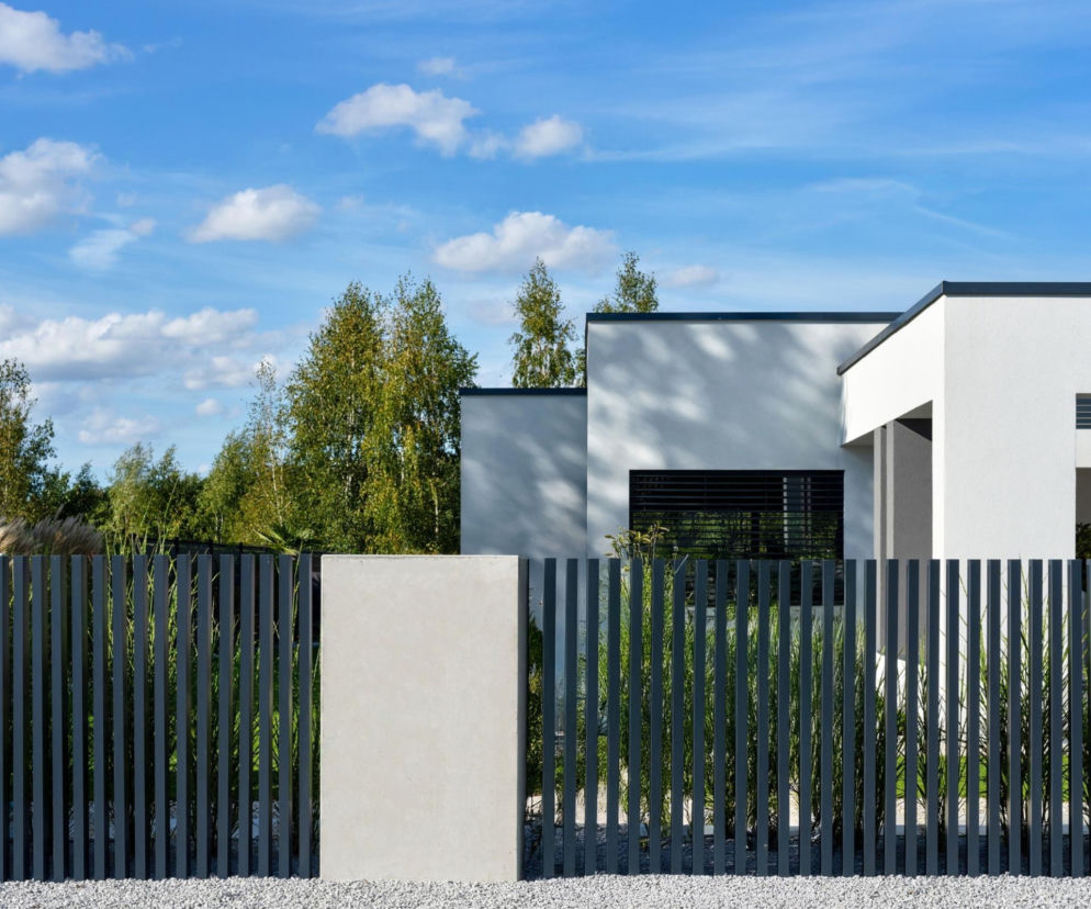Nowoczesne ogrodzenie palisadowe Wiśniowski w kolorze antracytowym, z betonowymi słupkami i podłożem ze żwiru, przed współczesnym domem z białej elewacji. Dowiedz się więcej o wyborze ogrodzenia na naszym portalu.