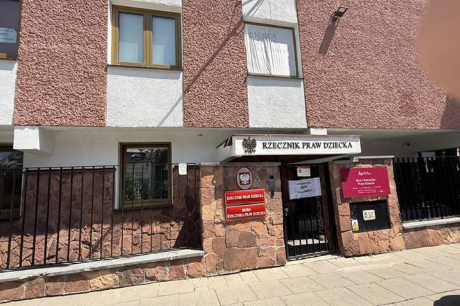 Fasada Biura Rzecznika Praw Dziecka z tablicami informacyjnymi i godłem Polski. Na ogrodzeniu widoczny transparent z napisem MURDZA RPD PRACOWNICY, co odnosi się do protestu i niezgody na medialne informacje. Więcej na ten temat przeczytasz na naszym portalu.