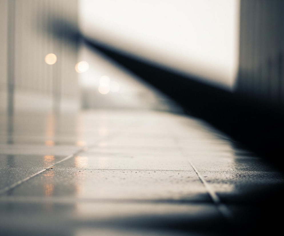 Ścieżka z płytek o szarym odcieniu, pokryta drobnymi kroplami wody, rozciąga się od dołu obrazu w kierunku centrum. Na jej powierzchni widoczne są jaśniejsze, rozmyte odbicia okrągłych źródeł światła. Po lewej stronie ścieżki znajduje się pionowy rząd ciemnych, niewyraźnych obiektów, a po prawej, w dolnej części, dominuje ciemna, niewyraźna bryła, tworząca ostry, diagonalny cień. Tło jest jasne i całkowicie rozmyte, z kilkoma okrągłymi punktami świetlnymi.