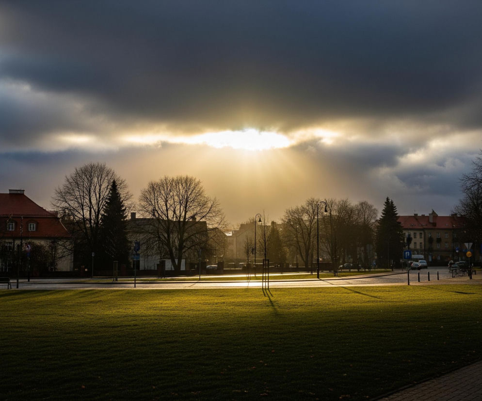 Osoba w ciemnej kurtce z kapturem i czapce, zwrócona plecami do obserwatora, idzie brukowanym chodnikiem po prawej stronie kadru. Po lewej rozpościera się szeroki, zielony trawnik, częściowo oświetlony promieniami słońca. Na drugim planie widoczne są ulicę i szereg budynków z dachami z czerwonej dachówki, częściowo zacienione, oraz liczne drzewa bez liści, rysujące się jako ciemne sylwetki na tle nieba. W centrum kadru słońce przebija się przez warstwę ciemnych chmur, tworząc jasny pas światła i rozchodzące się promienie, z lewej strony nieba widać fragmenty błękitu z białymi obłokami.
