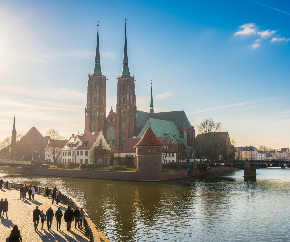 Na pierwszym planie, po lewej stronie, rozświetlona słońcem ścieżka spacerowa wzdłuż rzeki, po której idzie wiele osób. W tle dominuje duży, ceglany kościół z dwiema wysokimi, ostro zakończonymi wieżami pokrytymi zielonymi dachami oraz mniejszymi wieżyczkami. Rzeka z ciemnozieloną wodą otacza wyspę, na której stoi kościół i inne budynki, a w oddali widać kilka mostów z przechodzącymi ludźmi, oraz gęsto zabudowany brzeg z kamienicami. Po lewej stronie niebo jest jasne, niemal białe od słońca, przechodzące w błękit po prawej, z widocznymi smugami chmur.