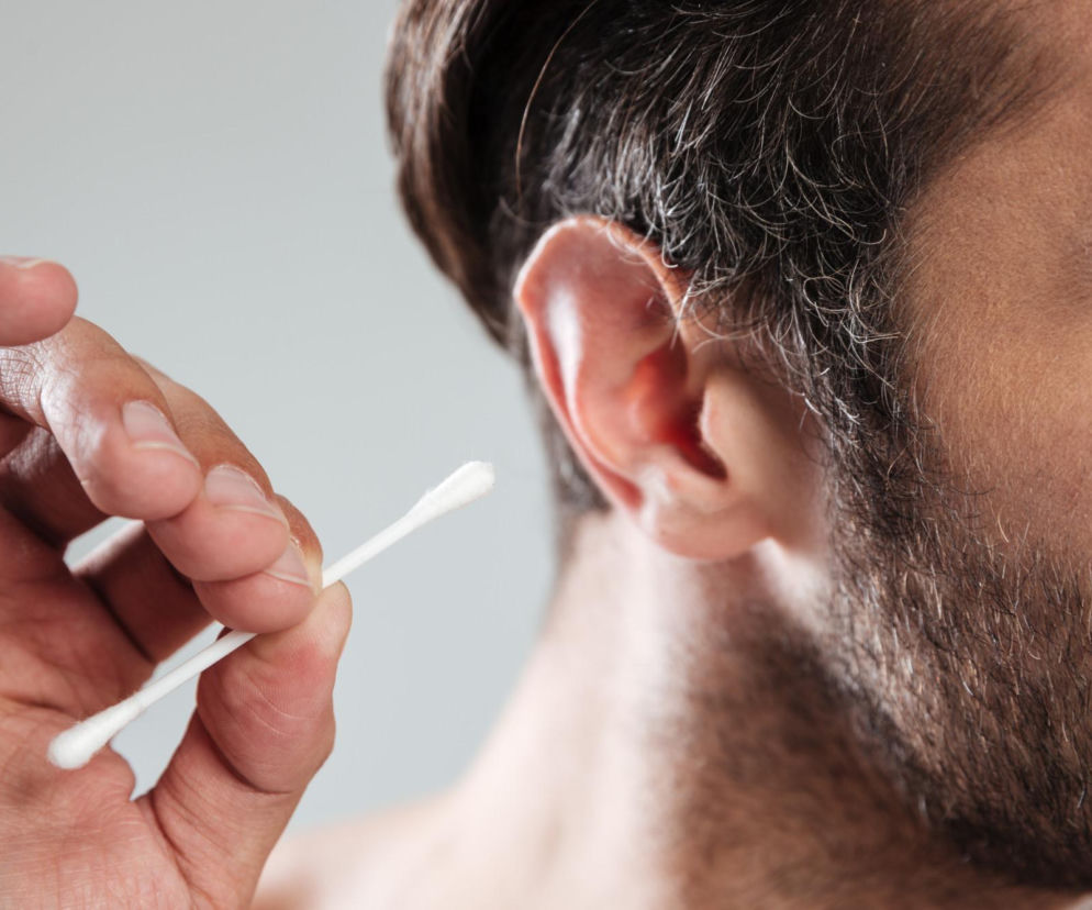 Mężczyzna z zarostem trzyma patyczek higieniczny przy uchu, co symbolizuje popularny, lecz niewłaściwy nawyk czyszczenia uszu. Na Poradnik Zdrowie znajdziesz wskazówki, jak prawidłowo osuszyć uszy, by uniknąć zapalenia zwanego uchem pływaka.