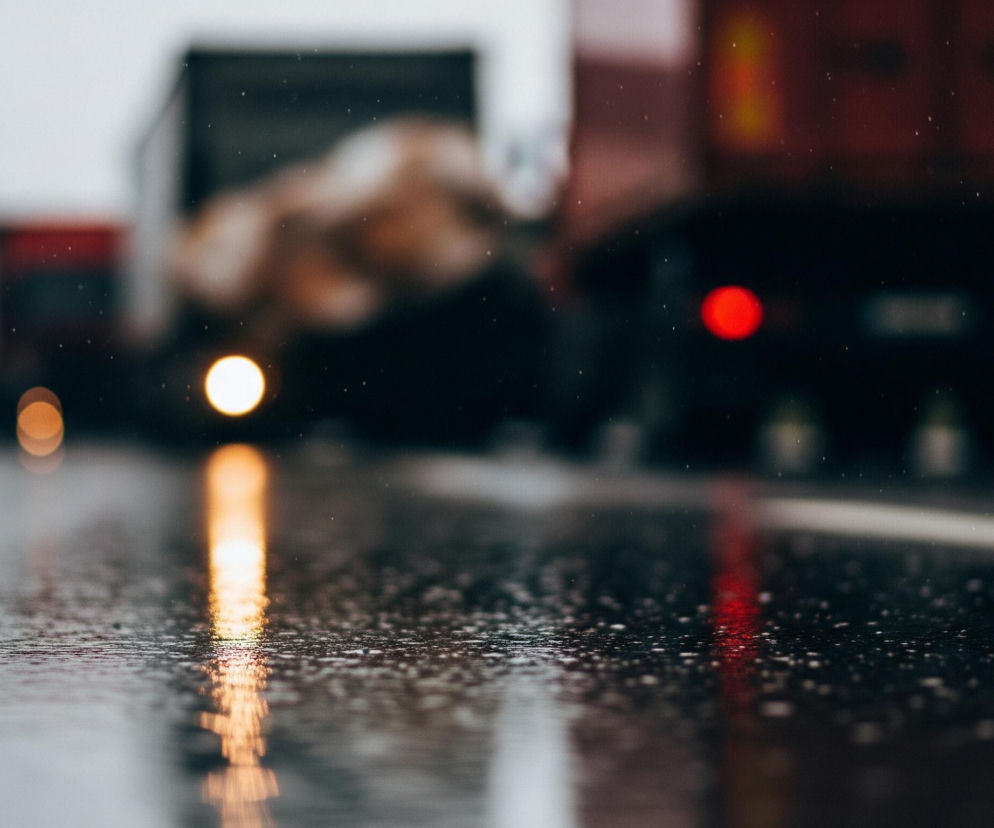 Na pierwszym planie, w dolnej części obrazu, widoczna jest mokra asfaltowa droga z licznymi kroplami wody tworzącymi drobne odbicia. Biała linia oddzielająca pasy ruchu jest rozmyta po lewej stronie. Na drodze odbijają się rozmyte światła pojazdów: pośrodku jasne, żółtopomarańczowe, a po prawej intensywnie czerwone. W tle, powyżej odbić, widać rozmyte sylwetki kilku ciężarówek lub innych dużych pojazdów, z czego jedna, po prawej stronie, ma czerwone elementy i wyraźnie widoczne okrągłe, czerwone światło tylne. Całość utrzymana jest w chłodnych, szaro-granatowych barwach z akcentami jaskrawego pomarańczu i czerwieni, sugerując pochmurny dzień lub zmierzch i mokrą nawierzchnię.