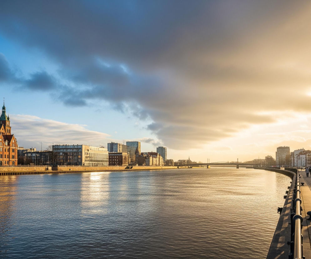 Rzeka zajmuje centralną część obrazu, odbijając złote światło zachodzącego słońca z prawej strony kadru oraz błękit nieba z lewej, tworząc wyraźną linię horyzontu. Na lewym brzegu widać ciąg budynków, wśród których wyróżnia się duży, ceglany obiekt z zielonym dachem i wieżą, natomiast na prawym brzegu, w bliższym planie, znajduje się kamienna promenada z balustradą, po której spacerują ludzie, rzucając długie cienie. W tle rozciąga się miejski krajobraz z mostem i dalszymi budynkami. Niebo jest częściowo zachmurzone, z ciemnymi chmurami po lewej stronie i jaśniejszymi, złotymi obłokami po prawej, gdzie słońce wyraźnie świeci.