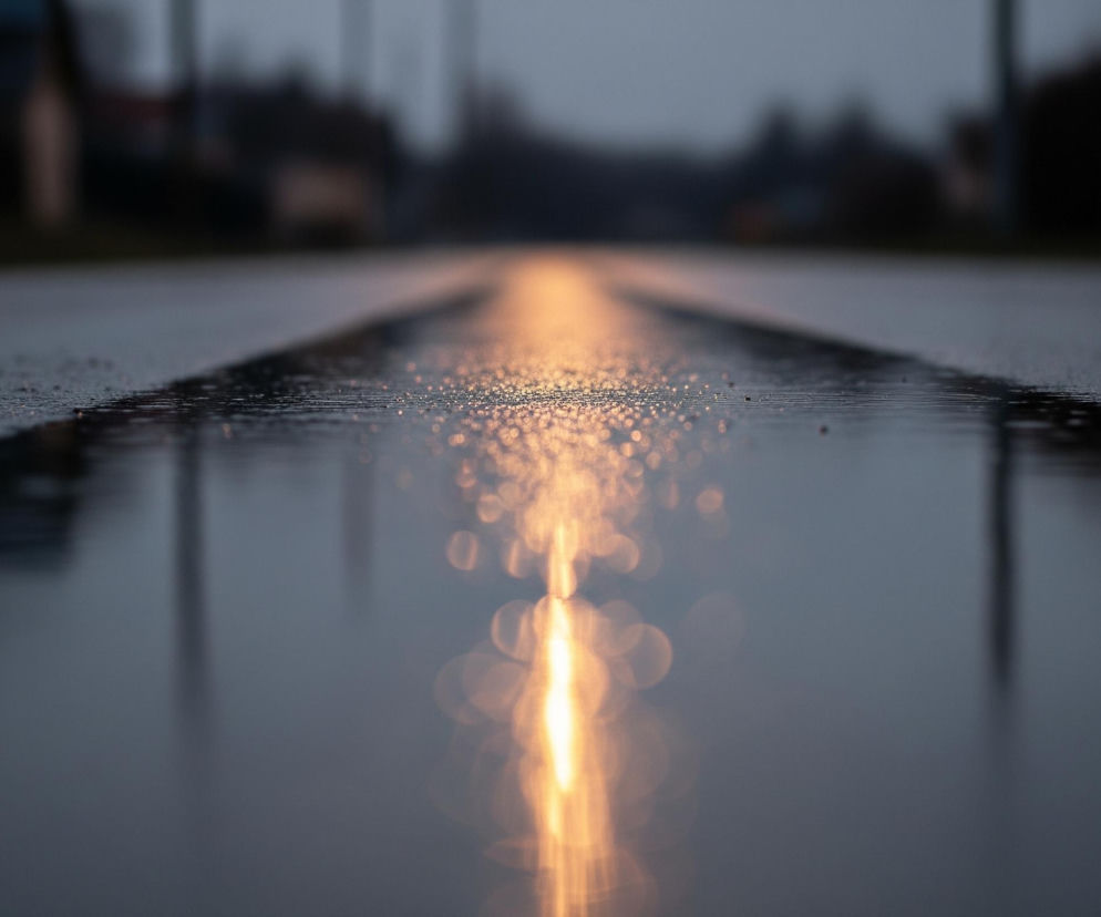 Mokra, asfaltowa droga biegnie przez centrum kadru, rozciągając się w dal. Na jej powierzchni widoczne są liczne krople wody, które tworzą błyszczące refleksy, zwłaszcza w środkowej części, gdzie intensywna, rozmyta smuga pomarańczowego światła odbija się od tafli wody. Lewa strona drogi i jej pobocze są ciemniejsze i bardziej matowe, z kilkoma drobnymi refleksami. Tło jest bardzo płytko nieostre, ukazując niewyraźne zarysy drzew i budynków w szaro-niebieskich tonacjach.