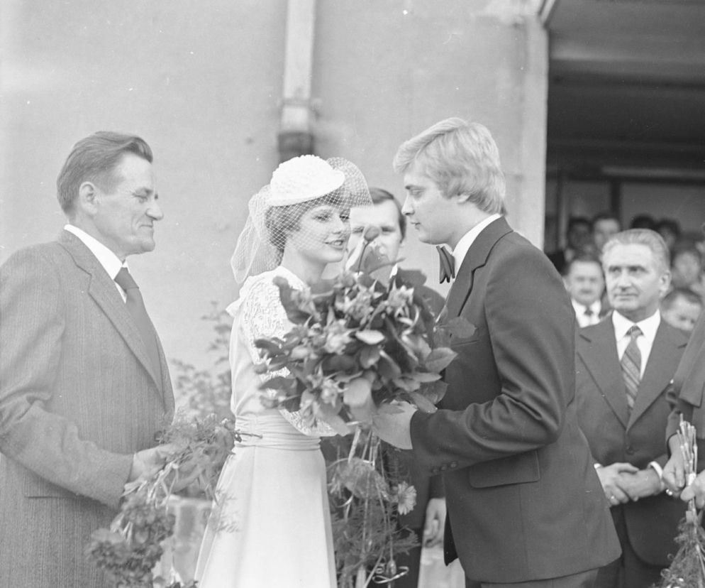 Uśmiechnięta młoda para w strojach ślubnych z bukietem kwiatów, otoczona gośćmi, w retro estetyce wesela z czasów PRL. Artykuł na naszym portalu przybliża klimat tamtych uroczystości i zachęca do rozwiązania quizu o weselach za komuny.