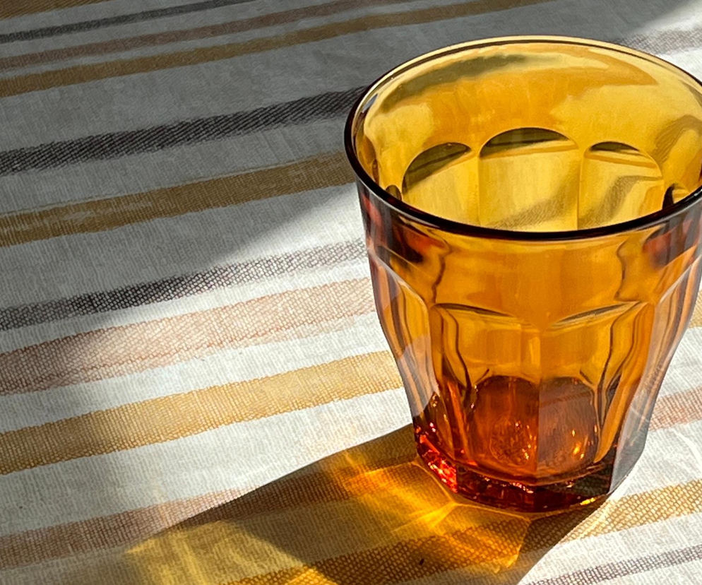 Bursztynowa szklanka Duralex o fasetowanych ściankach, stojąca na jasnym obrusie w paski, częściowo oświetlona promieniami słońca. Kultowa marka Duralex uratowana, o czym przeczytasz na Super Biznes.