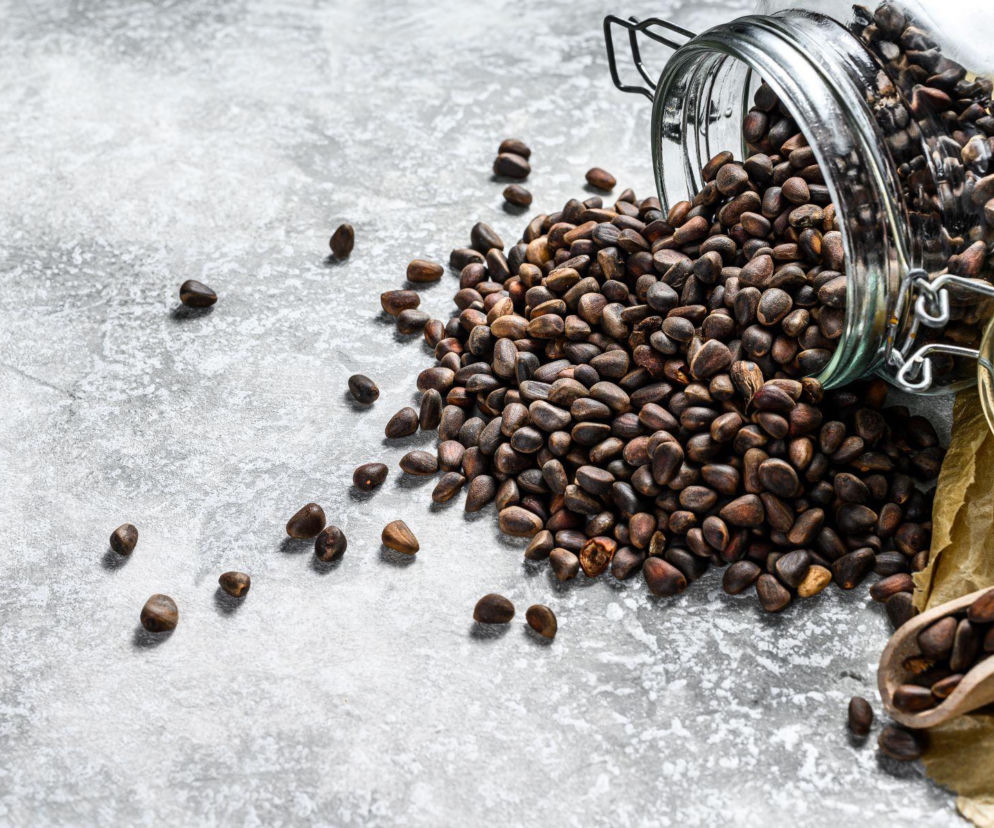Ziarna kawy wysypujące się ze szklanego słoika na szary, betonowy blat, obok papierowa torebka i drewniana łyżeczka. Artykuł na Poradnik Zdrowie wyjaśnia popularne mity o kawie i jej wpływie na nawodnienie organizmu.