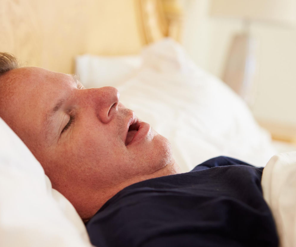 Mężczyzna w średnim wieku śpi w łóżku z otwartymi ustami, ubrany w granatową koszulkę. Leży na białej poduszce, a tło jest rozmyte, co tworzy spokojną atmosferę. Mówienie przez sen to temat, o którym przeczytasz więcej na Poradnik Zdrowie.