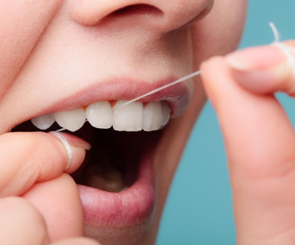 Uśmiechnięta osoba z nitką dentystyczną czyszcząca zęby techniką „C” w celu prawidłowej higieny jamy ustnej. Zdjęcie ilustruje, jak nitkować zęby, aby zmniejszyć krwawienie dziąseł, o czym można przeczytać na portalu Poradnik Zdrowie.