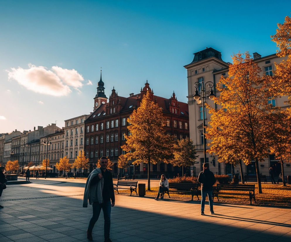 Miejski plac z dużą, ciemną fontanną po lewej stronie, z której tryskają strumienie wody. Brukowany plac rozciąga się w kierunku wysokich budynków w stylu europejskim, o różnobarwnych elewacjach, z których jeden wyróżnia się wieżą zegarową. Po prawej stronie placu biegnie trawnik z licznymi ławkami i pomarańczowo-żółtymi drzewami, których liście pokrywają również podłoże. Słońce, znajdujące się po lewej stronie kadru, rzuca długie cienie na bruk, oświetlając złocistym światłem korony drzew i budynki, podczas gdy niebieskie niebo z delikatnymi chmurami dominuje nad horyzontem.