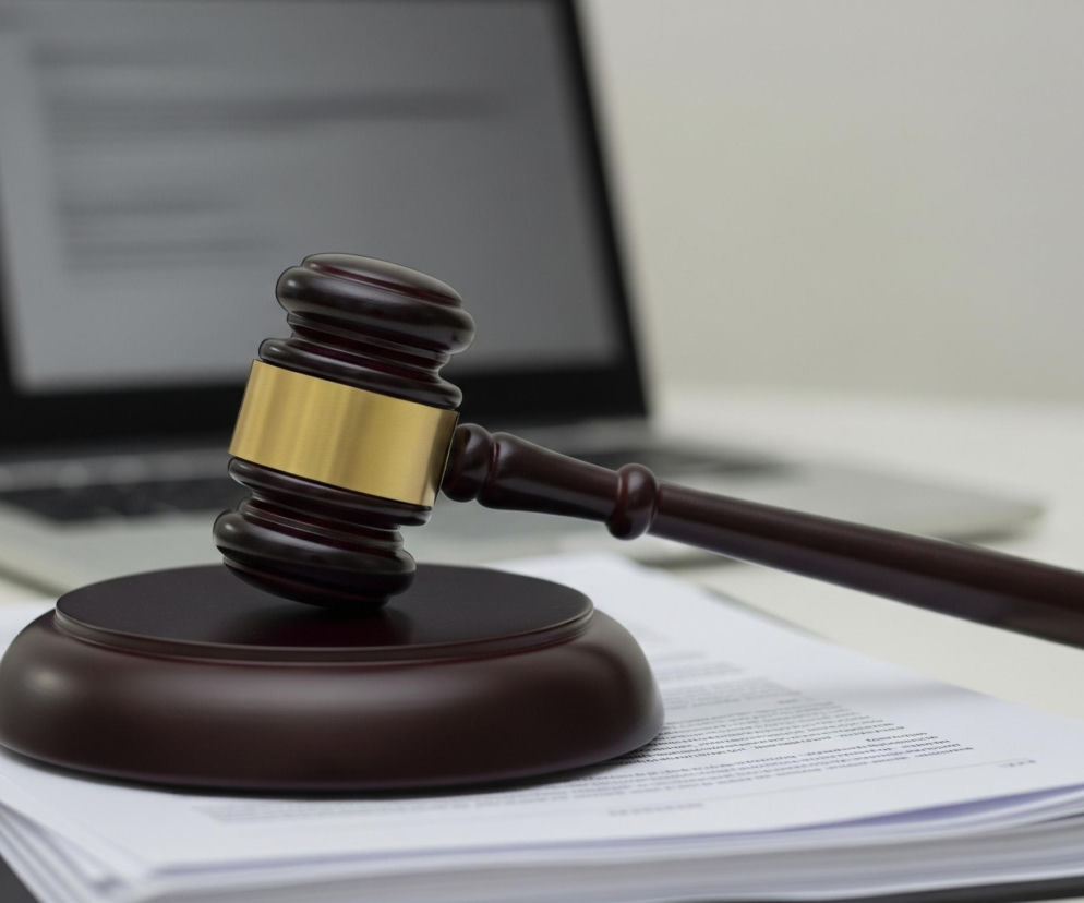 Na pierwszym planie, po prawej stronie, leży drewniany młotek sędziowski w kolorze ciemnego brązu, z metalową obręczą w odcieniu złota w górnej części. Jego rękojeść jest skierowana w prawo, natomiast główka spoczywa na okrągłej podstawce, również w ciemnobrązowym kolorze drewna. Poniżej młotka znajduje się stos białych kartek z widocznym na wierzchu drobnym czarnym tekstem, leżący na czarnej teczce. W tle, po lewej stronie, rozmyty jest otwarty laptop w srebrnym kolorze, z widocznym fragmentem ekranu prezentującym białą zawartość z ciemnym tekstem. Całość kompozycji utrzymana jest w stonowanych barwach.