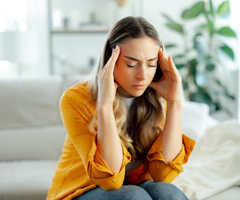 Młoda kobieta z długimi, blond włosami siedzi na kanapie, trzymając dłonie przy skroniach, z zamkniętymi oczami i cierpiącym wyrazem twarzy, co symbolizuje silny ból głowy. To ilustracja do artykułu Poradnika Zdrowie o objawach brain zaps i ich przyczynach.