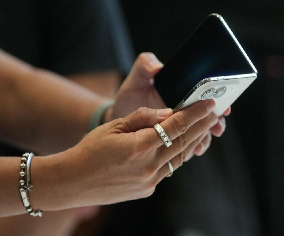 Dłonie osoby trzymającej na wpół złożony biały iPhone Duo z dwoma obiektywami. O nowym smartfonie Apple przeczytasz na Eska.