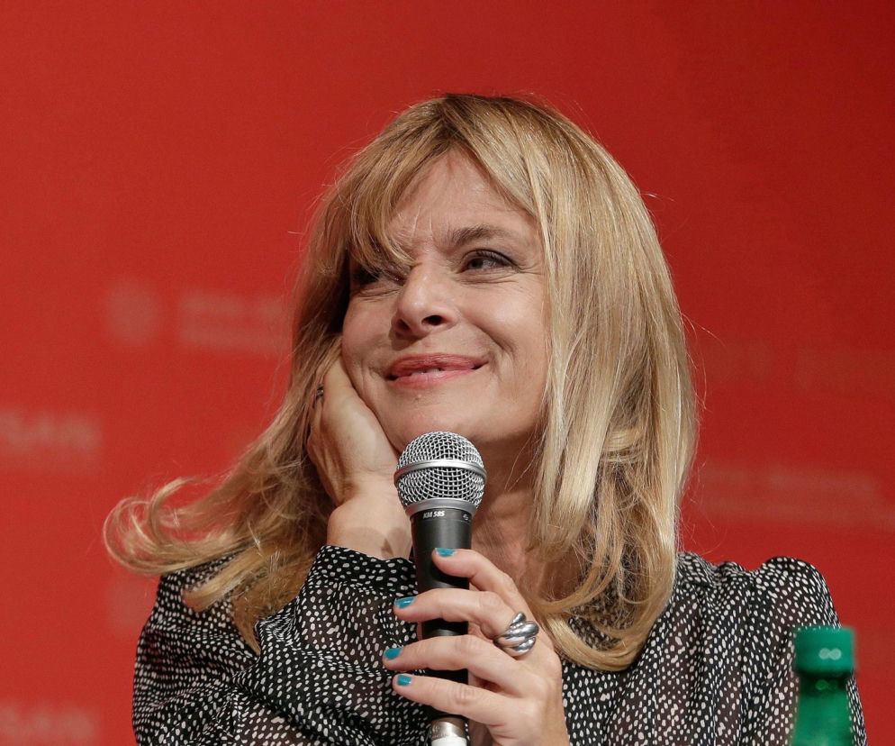 Portret uśmiechniętej Nastassji Kinski o blond włosach, trzymającej mikrofon i opierającej głowę na dłoni, podczas spotkania w Toruniu, gdzie odbierze Złotego Anioła. O kulisach wizyty przeczytasz więcej na naszym portalu.