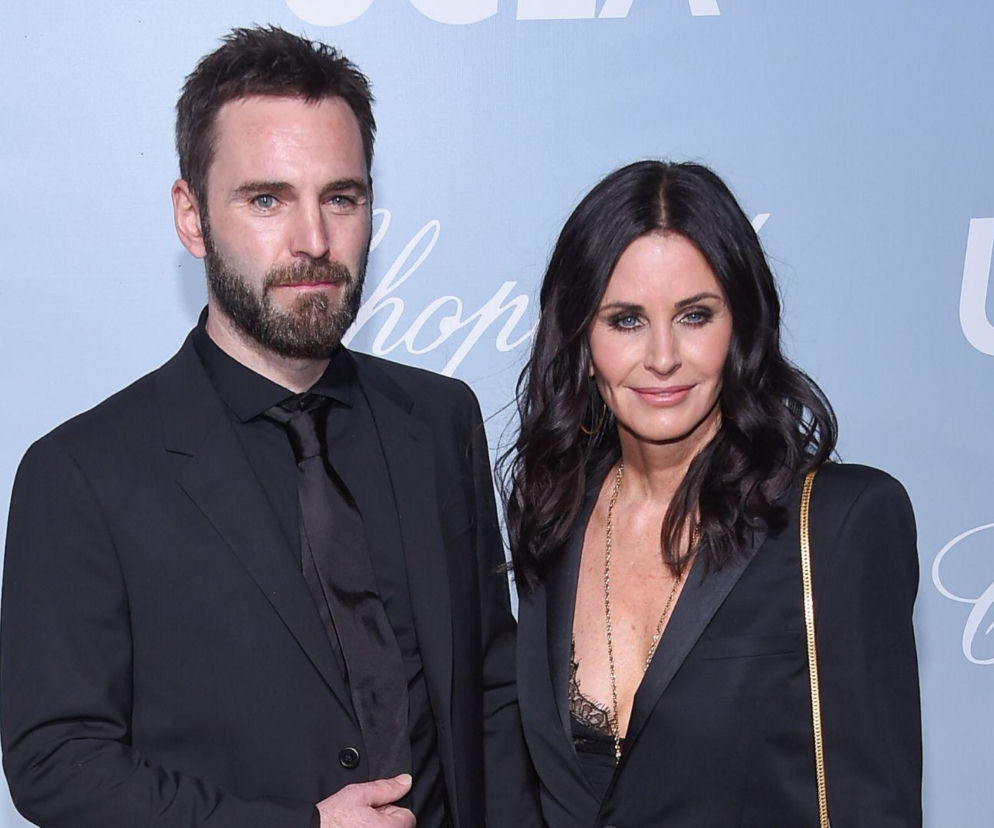 Courteney Cox i Johnny McDaid, ubrani w eleganckie czarne stroje, uśmiechają się na tle jasnego baneru z napisem UCLA i Chopard. Para pozuje na uroczystej imprezie, co nawiązuje do burzliwej historii ich związku, o której przeczytasz na naszym portalu.