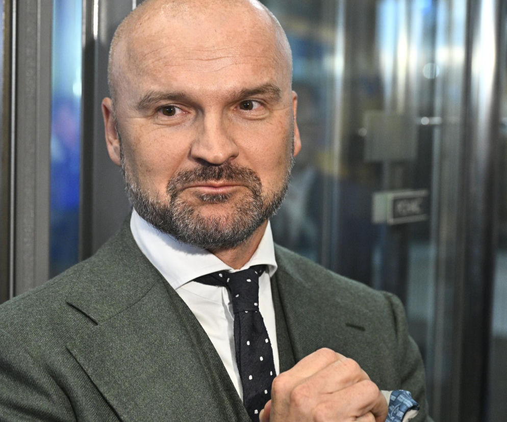 Rafał Brzoska, prezes InPostu, w zielonym garniturze, białej koszuli i krawacie w białe grochy, z zaciskającymi się dłońmi. W tle widać fragmenty przeszklonych ścian. O jego opinii na temat polskiego biznesu i podatków przeczytasz na Super Biznes.