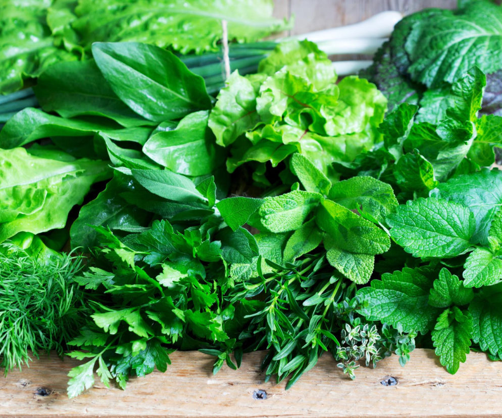 Zbiór świeżych zielonych warzyw liściastych, takich jak sałata, szpinak, jarmuż, pietruszka, koperek, mięta i szczypiorek, leżących na drewnianym stole. Zielone warzywa w diecie pomagają obniżyć ciśnienie, więcej o tym na Poradnik Zdrowie.