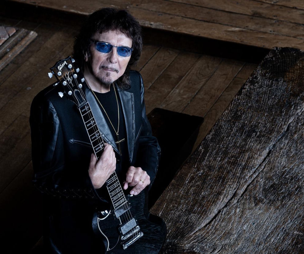 Gitarzysta Tony Iommi w ciemnej marynarce i niebieskich okularach trzyma czarną gitarę Gibson. O nowej płycie czytaj w EskaRock.