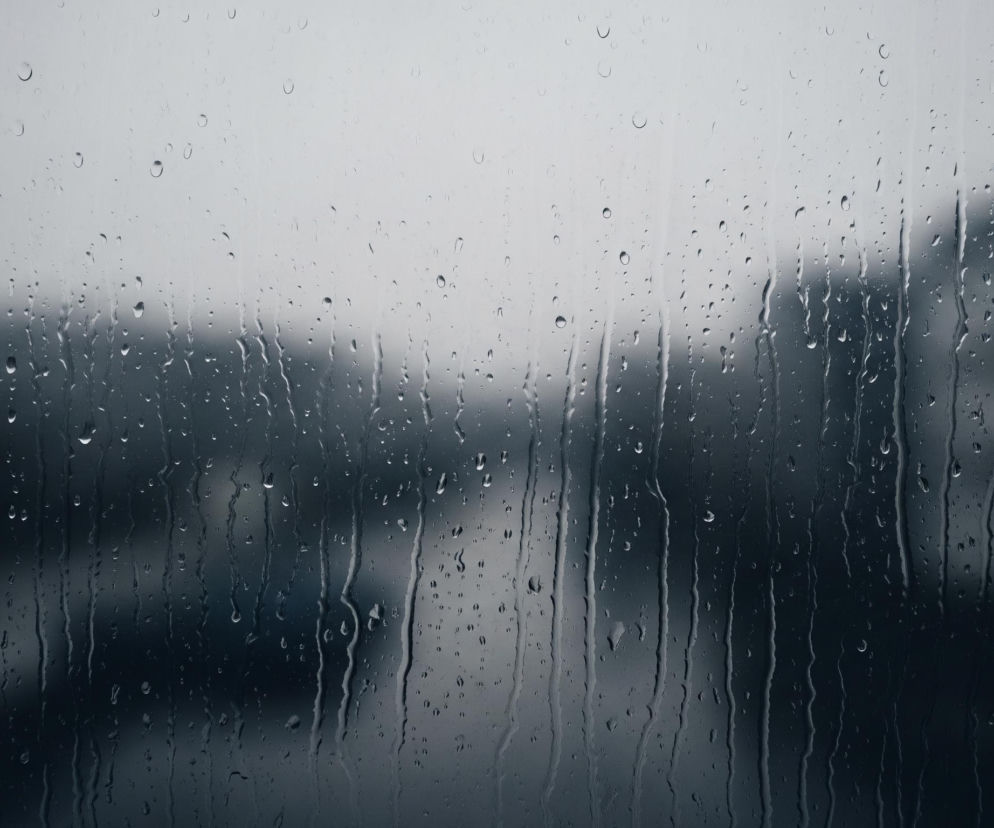 Przednia płaszczyzna obrazu to szyba pokryta licznymi kroplami deszczu o różnej wielkości, w tym smugami spływającej wody. Tło jest rozmyte i niewyraźne, z widocznymi odcieniami szarości i ciemnego granatu, sugerującymi sylwetki budynków lub drzew. Górna część obrazu jest jaśniejsza, przyjmując kolor jasnoszary, natomiast dolna część jest znacznie ciemniejsza, z przewagą ciemnych, stonowanych barw.