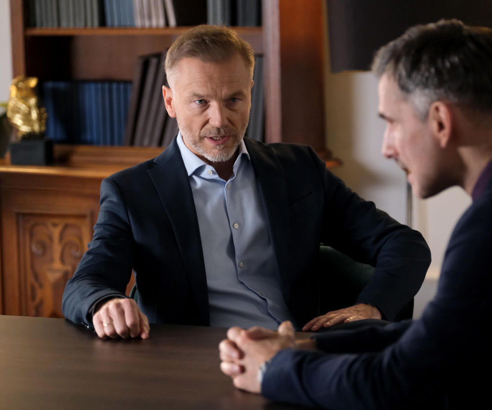 Andrzej Budzyński (Krystian Wieczorek) z serialu M jak miłość, w granatowej marynarce i błękitnej koszuli, siedzi przy stole i rozmawia z Kamilem Grycem (Marcin Bosak), widocznym z profilu. Budzyński ma poważną minę, patrzy prosto w kamerę. O tej i innych historiach przeczytasz na Super Seriale.