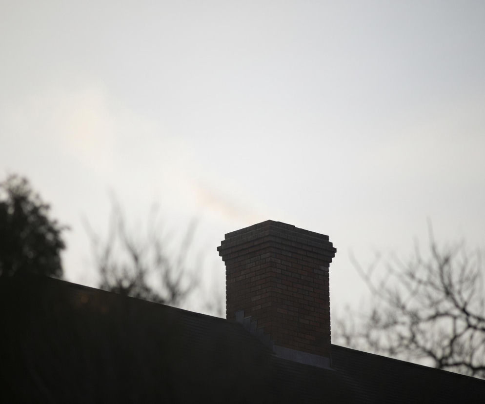 Centralnie na obrazie dominuje ceglany komin w odcieniach brązu, wznoszący się z ciemnego dachu. Nad kominem unosi się delikatna smuga dymu w kolorze szarym, niemal niewidoczna na jasnym, rozświetlonym tle nieba. W górnej części zdjęcia, na tle jasnego nieba, widoczne są rozmyte, ciemne sylwetki drzew, z widocznymi gałęziami po prawej stronie i gęstszą koroną po lewej.