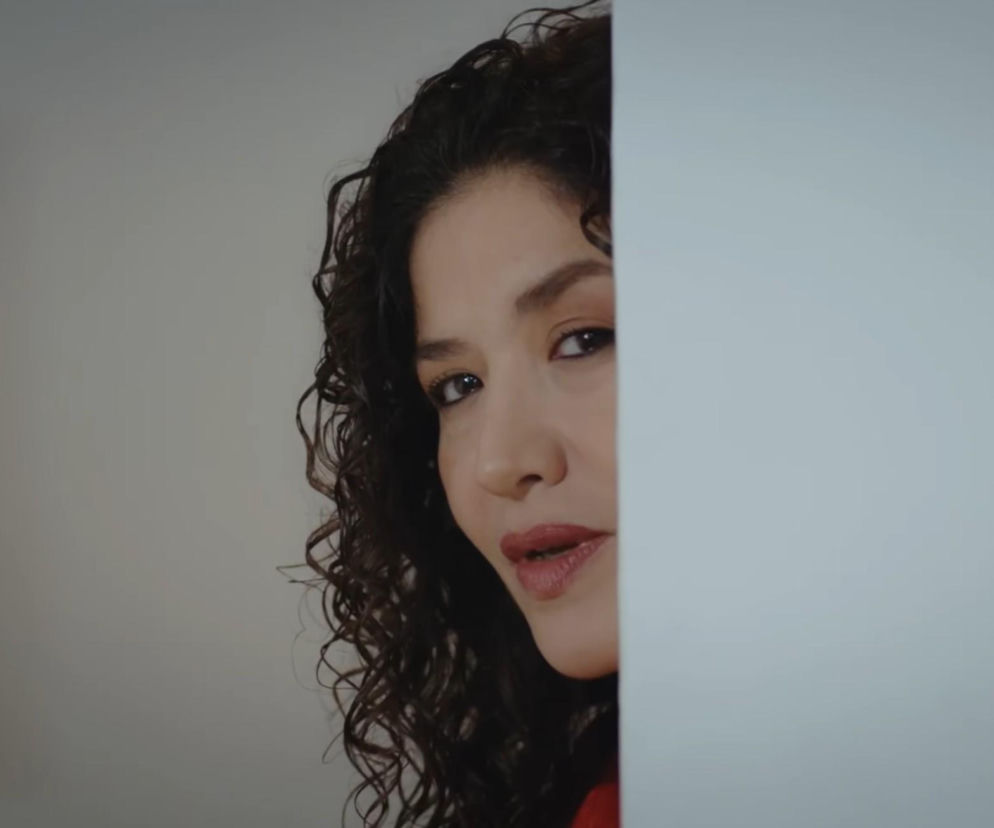 Kobieta z kręconymi włosami i ciemnym makijażem oczu, wyglądająca zza białej ściany, co symbolizuje jej ukryte intencje. Postać Cansel z serialu Dziedzictwo, realizująca swój plan przejęcia majątku Kyrymlych, o czym przeczytasz na Super Seriale.