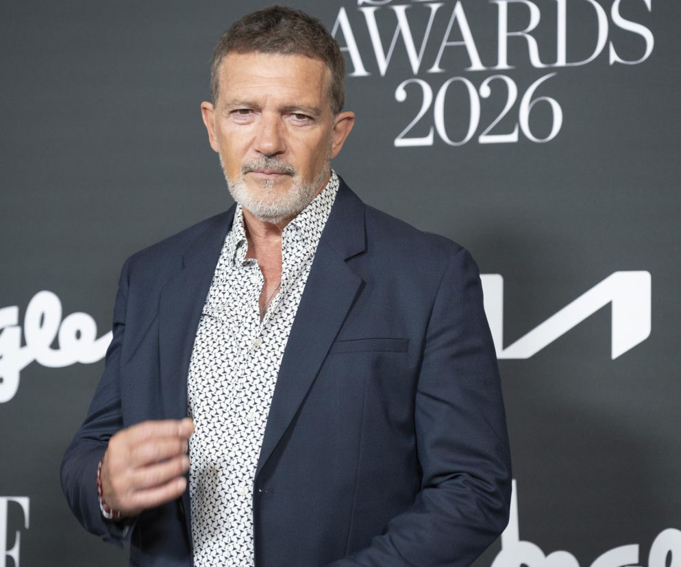 Aktor Antonio Banderas w eleganckiej, granatowej marynarce i białej koszuli w ciemny wzór, z lekko rozpiętym kołnierzykiem, patrzy prosto w obiektyw. Na tle widoczne są fragmenty logotypów „AWARDS 2026” i innych, niewyraźnych napisów. Czytaj więcej o śledztwie na naszym portalu.