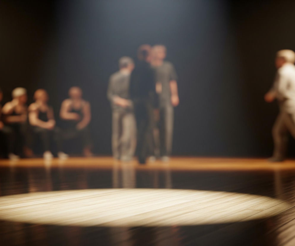 Drewniana scena jest oświetlona silnym, okrągłym snopem światła na pierwszym planie, z delikatnymi odbiciami na błyszczącej powierzchni. W tle, rozmyte postacie ludzi są widoczne na ciemnym tle, z kilkoma osobami stojącymi i kilkoma siedzącymi po lewej stronie. Jeden człowiek w jasnym ubraniu, również rozmazany, zmierza w prawą stronę obrazu, oddalając się od pozostałych.