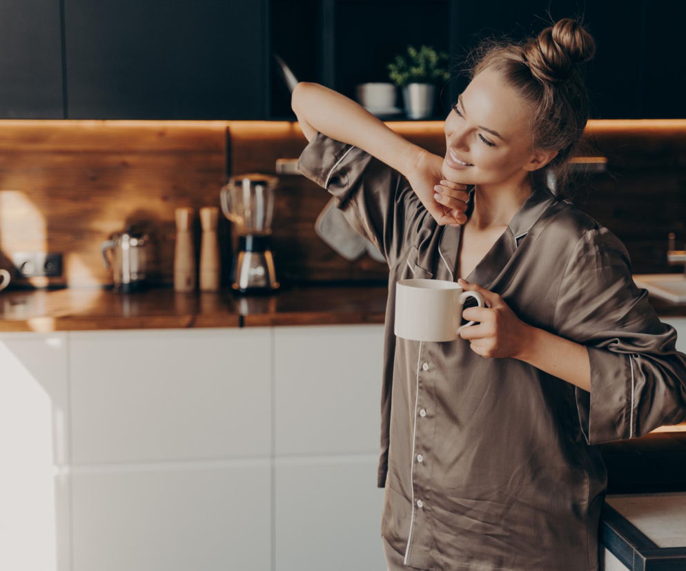 Młoda kobieta w piżamie przeciąga się z kubkiem kawy w ręku, uśmiechając się, stojąc w słonecznej kuchni o poranku. Dowiedz się, dlaczego poranna kawa zaraz po przebudzeniu może odbierać energię, w artykule na Poradnik Zdrowie.