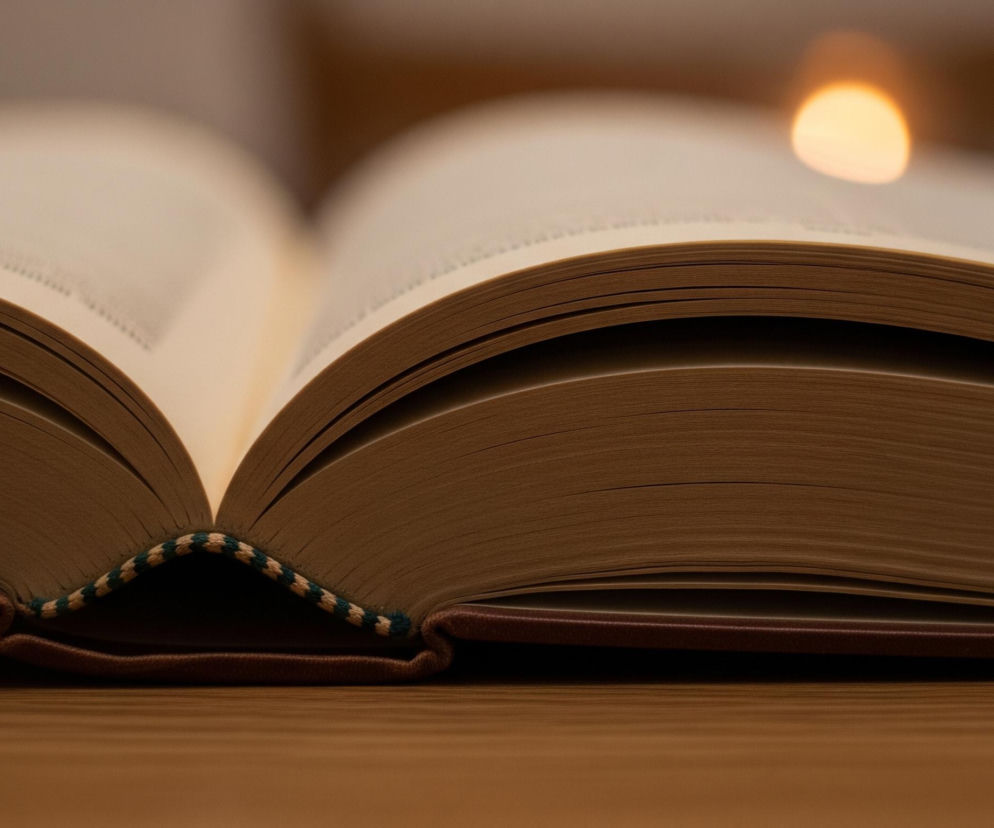 Otwarta książka z brązową okładką spoczywa na drewnianej, brązowej powierzchni. Jej strony mają lekko beżowy odcień i są zagięte w łagodny łuk, tworząc zagłębienie w środku, gdzie widoczna jest ozdobna, pleciona opaska w zielono-beżowe paski. W prawym górnym rogu obrazu, w tle, znajduje się rozmyte, jasne pomarańczowe światło. Całość skąpana jest w ciepłym, przytłumionym oświetleniu.
