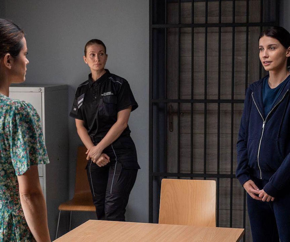 Trzy kobiety w pomieszczeniu przypominającym komisariat lub pokój przesłuchań: Kasia, w kwiecistej sukience, stoi tyłem do widza. Obok strażniczka więzienna w czarnym mundurze i Justyna w dresie, z kratami więziennymi w tle. Scena z serialu M jak miłość o problemach Kasi z powodu siostry kryminalistki, co można śledzić na Super Seriale.