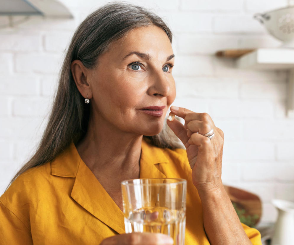 Starsza kobieta w musztardowej koszuli z siwymi włosami, trzymająca w dłoni szklankę wody i przezroczystą kapsułkę suplementu potasu. Analizujemy, kiedy suplementacja potasu staje się groźna. Więcej na temat hiperkaliemii przeczytasz na portalu Poradnik Zdrowie.