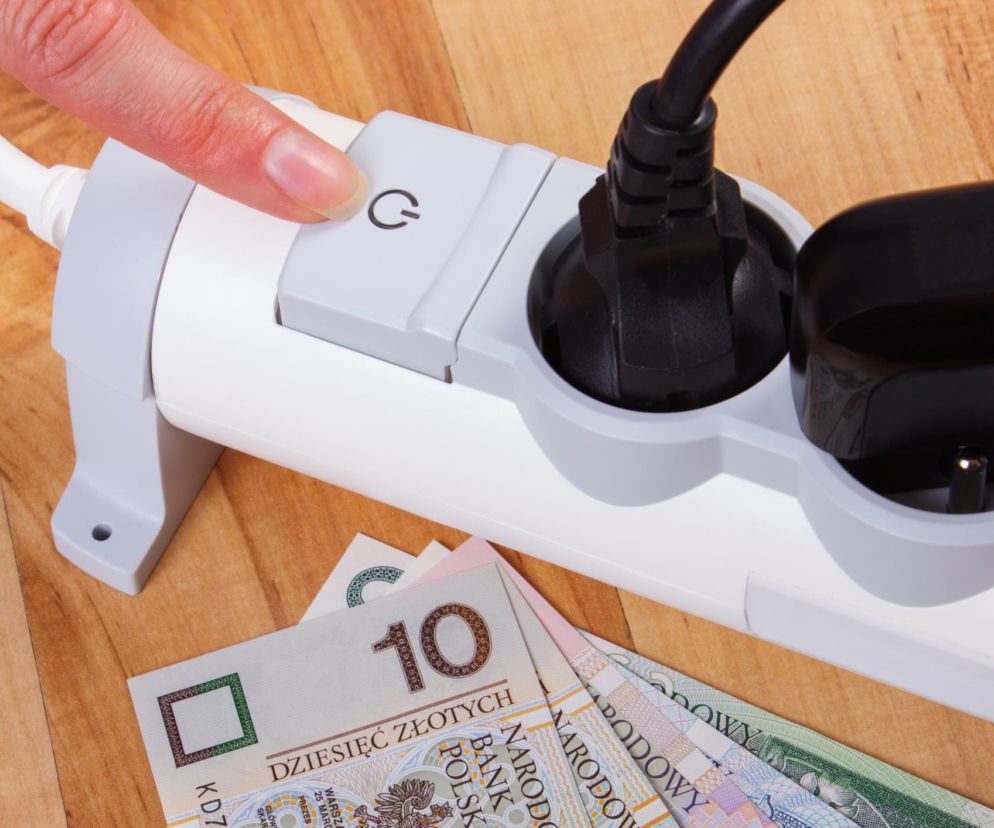 Ręka wciskająca przycisk włącznika zasilania w białej listwie zasilającej z trzema wtyczkami, symbolizująca oszczędzanie energii elektrycznej. Poniżej banknoty 10 zł i 20 zł, obrazując koszty zużycia prądu, o których więcej na naszym portalu.
