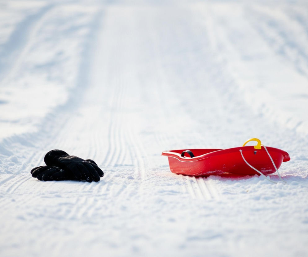 Na zaśnieżonym, lekko pagórkowatym terenie leżą czerwone sanki i dwie czarne rękawiczki. Sanki są widoczne po prawej stronie kadru, z żółtym uchwytem i białą linką, a obok nich, na środku obrazu, leżą złożone rękawiczki. Za sankami i rękawiczkami ciągną się trzy równoległe ślady na śniegu, prowadzące w głąb obrazu, gdzie teren jest niewyraźny i rozmyty. Całość skąpana jest w jasnym, zimowym świetle, tworzącym subtelne cienie i podkreślającym teksturę śniegu.