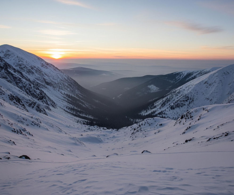 Ośnieżone góry rozciągają się, tworząc głęboką dolinę, w której dnie widać ciemne drzewa. Na tle nieba o zachodzie słońca, słońce, wschodzące lub zachodzące, maluje horyzont pomarańczowymi i różowymi odcieniami. Zbocza gór, pokryte warstwą śniegu, przechodzą od jasnych, podświetlonych obszarów do głębokich cieni w dolnych partiach, eksponując skaliste fragmenty. W oddali, pasma górskie zanikają w mglistej, niebieskiej poświacie, tworząc wrażenie głębi i rozległości krajobrazu.