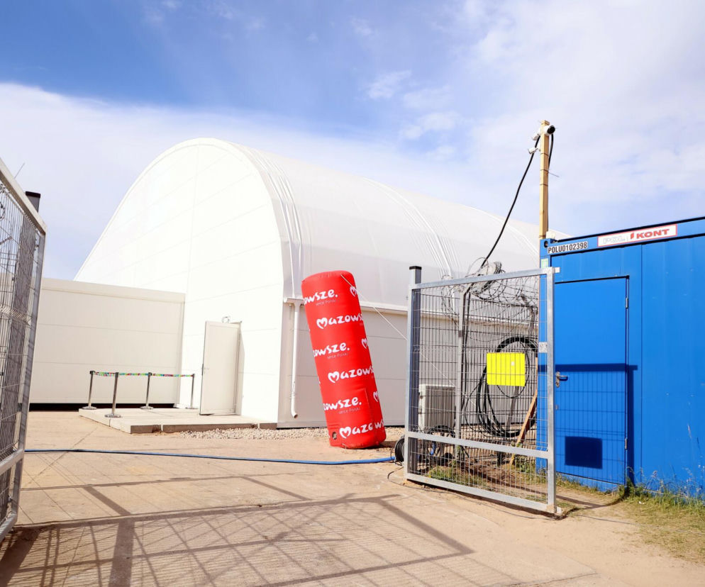 Zewnętrzny widok nowego terminala pasażerskiego na Lotnisku Warszawa-Modlin, przypominającego hangar, z białą konstrukcją i ogrodzeniem, symbolizujący rozwój i innowacyjne rozwiązania, o których można przeczytać na naszym portalu. Obok widoczny dmuchany słup z napisem Mazowsze. Serce Polski oraz niebieski kontener.