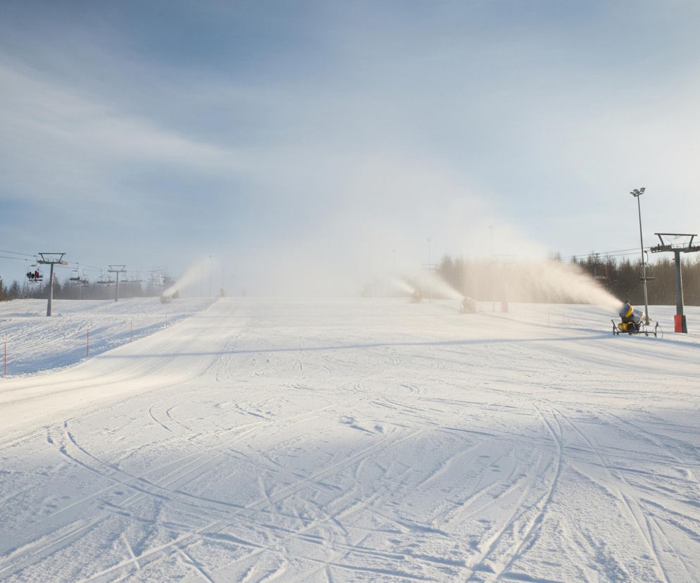 Ośnieżony stok narciarski rozciąga się w głąb obrazu, pokryty śladami od nart i ratraka. W oddali, po prawej stronie, żółta armatka śnieżna intensywnie produkuje biały śnieg, tworząc gęstą mgłę unoszącą się nad stokiem. W tle widoczna jest linia drzew oraz kilka słupów wyciągu krzesełkowego, na których zawieszone są krzesełka, niektóre z nich z pasażerami. Jasne, częściowo zachmurzone niebo dominuje górną część kadru, z delikatnymi smugami chmur.