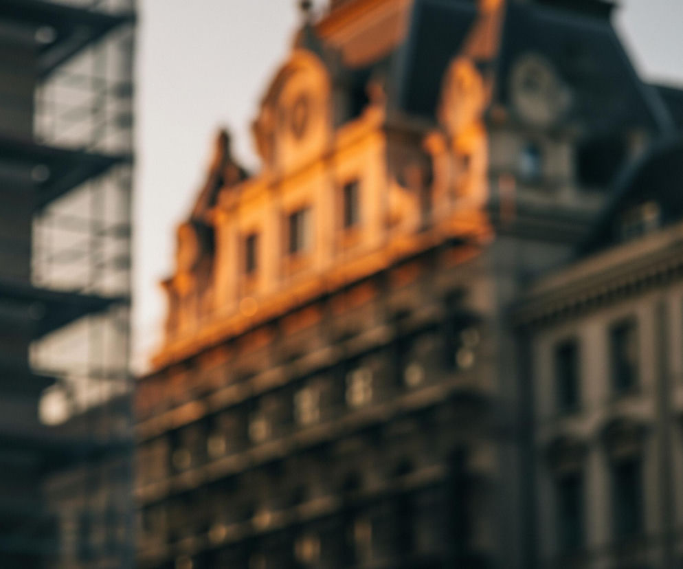 Głównym elementem obrazu jest rozmyty, neoklasycystyczny budynek w centrum, którego górne kondygnacje i mansardowy dach są intensywnie oświetlone ciepłym, pomarańczowo-złotym światłem zachodzącego słońca. Po lewej stronie widać fragment innej, ciemnej konstrukcji, przypominającej rusztowanie lub nowocześniejszy budynek, również nieostry. Po prawej stronie znajduje się kolejna, rozmyta fasada budynku w odcieniach szarości, z wyraźnymi oknami i elementami architektonicznymi. Całość kompozycji, wykonanej z płytką głębią ostrości, utrzymana jest w stonowanych barwach z dominującym pomarańczowym blaskiem na środku.