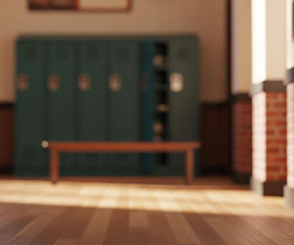 Na pierwszym planie, centralnie, rozciąga się podłoga z brązowych desek, częściowo rozjaśniona przez wpadające światło. W tle, na środku, znajduje się długie, wąskie drewniane ławka w kolorze pomarańczowo-brązowym. Za ławką widoczne są szeregi metalowych szafek w odcieniu ciemnej zieleni, z których jedna po prawej stronie jest otwarta. Po lewej stronie zdjęcia widać fragment ciemnobrązowych drzwi z dwoma jasnymi, prostokątnymi przeszkleniami. Po prawej stronie znajdują się ceglane filary, częściowo ukryte w cieniu, z fragmentem otwartych brązowych drzwi po skrajnej prawej stronie.