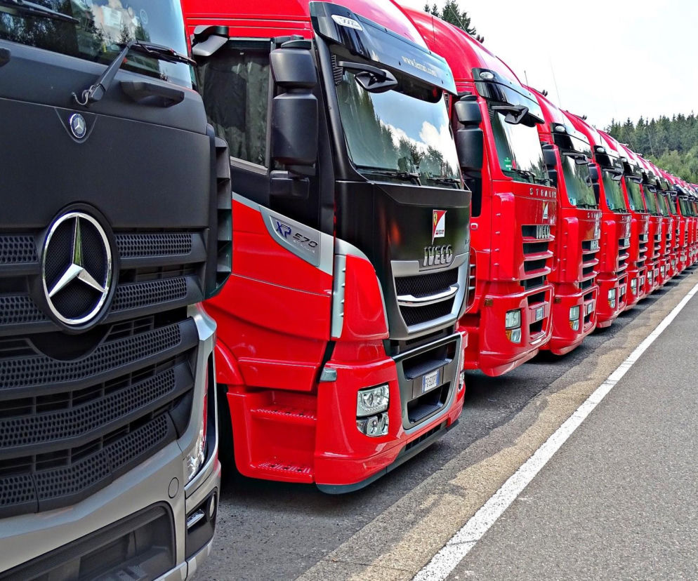 Rząd ciężarówek Mercedes i Iveco z czerwonymi kabinami zaparkowanych w rzędzie. Zdjęcie ilustruje temat dopłat do wymiany tachografów dla firm transportowych, o których przeczytasz więcej na portalu Super Biznes.