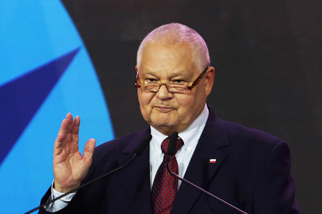 Prezes NBP Adam Glapiński w okularach, z białą koszulą i krawatem, w ciemnym garniturze z polską flagą na klapie, przemawia przy mównicy. Za nim widoczne jest tło w kolorach niebieskim i czarnym. O planach NBP i osobie Adama Glapińskiego przeczytasz na Super Biznes.