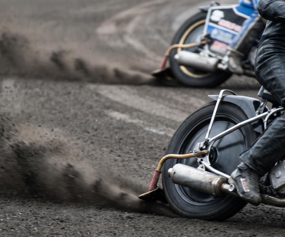 Żużlowiec w czarnym stroju w akcji na torze, ślizgający się na motocyklu żużlowym, z piachem unoszącym się za tylnym kołem. W tle drugi zawodnik na torze. Więcej o zwycięstwie Bartosza Zmarzlika we Wrocławiu przeczytasz na naszym portalu.