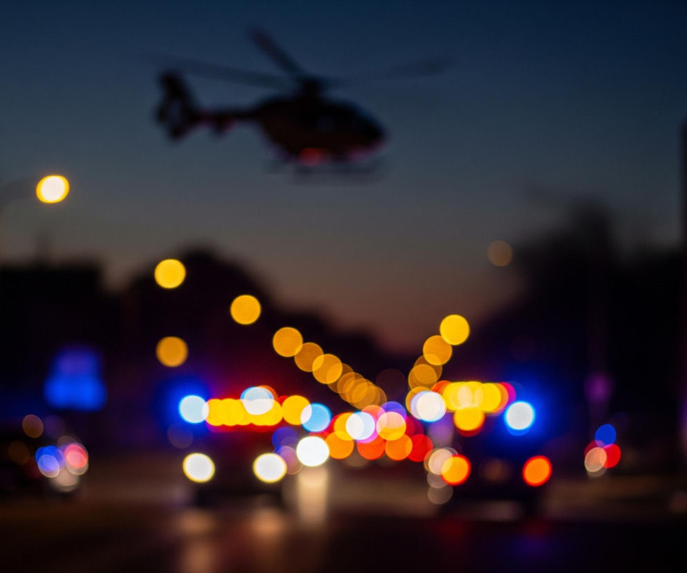 W centrum kadru rozmyty helikopter unosi się na tle ciemniejącego nieba, podświetlony od dołu przez czerwone światła. Poniżej, na poziomie ulicy, widoczne są rozmyte światła samochodów, głównie niebieskie, czerwone i białe, w otoczeniu licznych żółtych i pomarańczowych plam światła ulicznych latarni. Cała scena jest niewyraźna, tworząc efekt bokeh, z ciemnymi sylwetkami drzew i budynków po bokach.