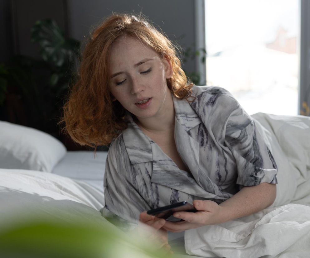 Kobieta o rudych, kręconych włosach, leżąca w łóżku i trzymająca smartfon, symbolizująca poranny nawyk, który może negatywnie wpływać na koncentrację. Więcej o wpływie telefonu na mózg przeczytasz na Poradnik Zdrowie.