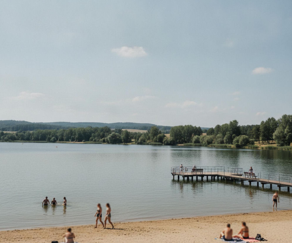 Osoby odpoczywające na piaszczystej plaży nad jeziorem z pomostem. O kąpieliskach w Małopolsce przeczytasz w serwisie Demo.