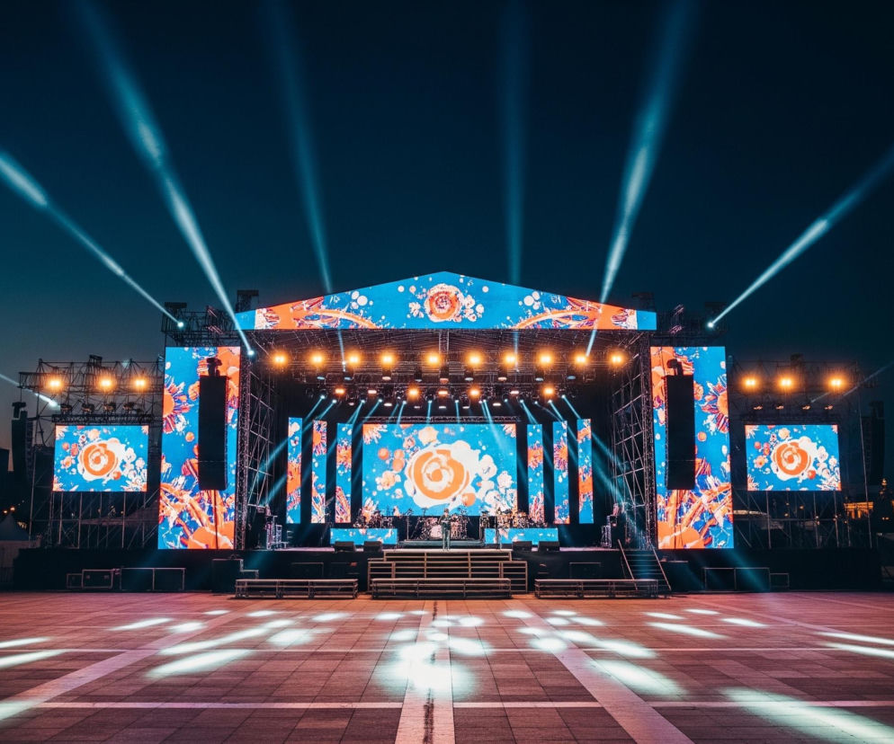 Przedstawiona jest szeroka perspektywa dużej, oświetlonej sceny koncertowej na zewnątrz, stojącej na otwartym placu po zmierzchu. Scena ma złożoną konstrukcję kratownicową z wieloma ekranami LED wyświetlającymi dynamiczne grafiki w kolorach niebieskim, pomarańczowym i białym, przypominające abstrakcyjne kwiaty i plamy farby. Liczne reflektory emitują jasne, niebieskawobiałe smugi światła w ciemne niebo, a także oświetlają przód sceny i plac. Na scenie widoczne są platformy dla instrumentów i muzyków, a także sprzęt nagłaśniający. Pusty, wybrukowany plac przed sceną odbija światło, ukazując rzędy białych barier ochronnych po lewej i prawej stronie. W tle, po bokach sceny, majaczą ciemne sylwetki budynków, z niektórymi oknami świecącymi żółtym światłem.