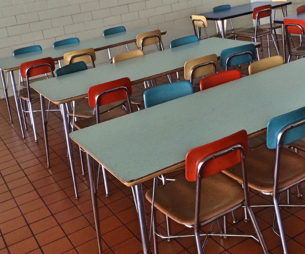 Szereg stolików i krzeseł w szkolnej stołówce z białymi kafelkami na ścianie, symbolizujący nadchodzące zmiany w żywieniu. Nowe regulacje stworzą stabilny rynek zbytu dla rolników na warzywa i rośliny strączkowe, o czym przeczytasz na Super Biznes.