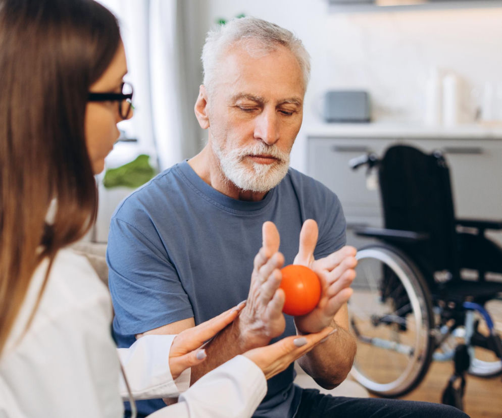 Fizjoterapeutka pomaga starszemu mężczyźnie z siwą brodą w ćwiczeniach dłoni z pomarańczową piłką, co wspiera rehabilitację po udarze. W tle widoczny wózek inwalidzki. Na portalu Poradnik Zdrowie znajdziesz informacje o tym, jak odzyskać sprawność.